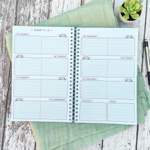 May include: A spiral-bound weekly planner open to a two-page spread for August 15-21. The white pages feature light blue horizontal lines, checkboxes, and small cartoon cat illustrations. Each day is labeled with the date and day of the week.