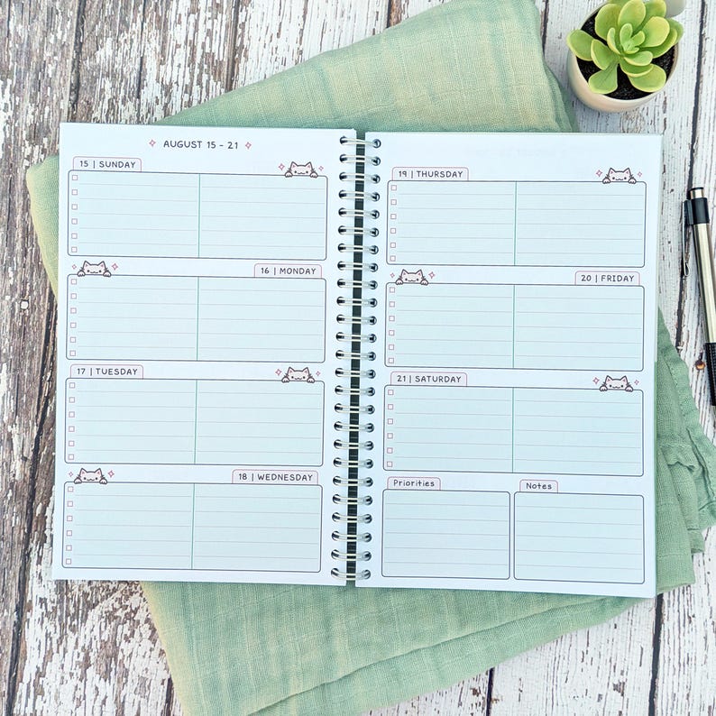 May include: A spiral-bound weekly planner open to a two-page spread for August 15-21. The white pages feature light blue horizontal lines, checkboxes, and small cartoon cat illustrations. Each day is labeled with the date and day of the week.