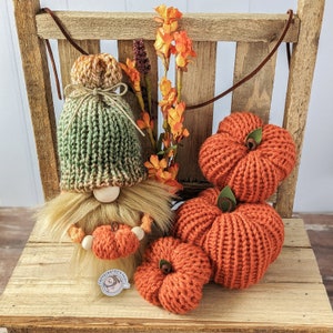 May include: A green and brown knitted gnome doll holding a small orange knitted pumpkin. The gnome is sitting on a wooden bench with three orange knitted pumpkins.