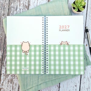 May include: An open 2027 planner with a white and light green gingham pattern on the bottom half of the pages. The planner features small cartoon cat illustrations, the text 2027 PLANNER, and a small catwillow studio logo on the bottom left page.