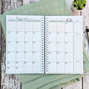 May include: A spiral-bound monthly planner open to August 2026. The calendar grid features light green headers for days of the week, numbered dates, and a small cat illustration. The right side includes sections for Goals, To-do lists, and Notes.