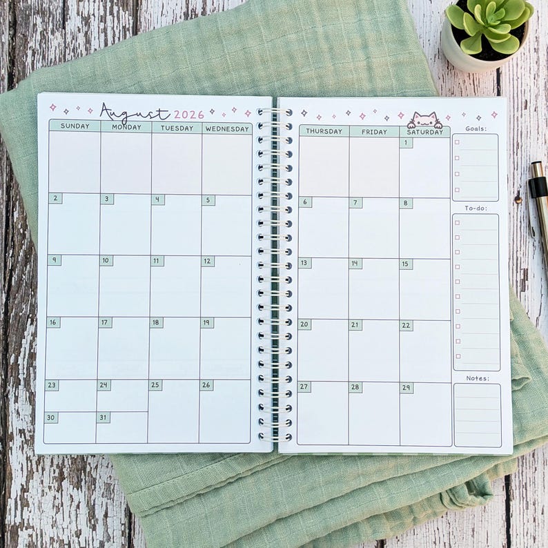 May include: A spiral-bound monthly planner open to August 2026. The calendar grid features light green headers for days of the week, numbered dates, and a small cat illustration. The right side includes sections for Goals, To-do lists, and Notes.
