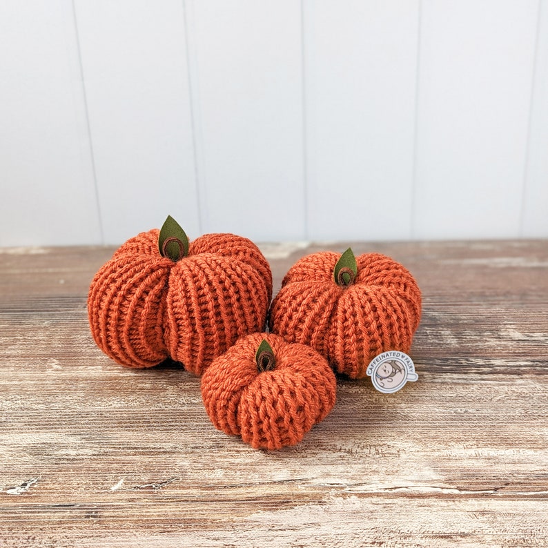 May include: Three knitted pumpkins in various sizes, the largest is orange and the smallest is a deep orange. The pumpkins are on a wooden surface.