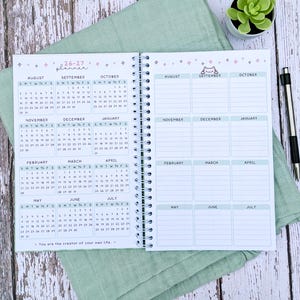 May include: A spiral-bound 26-27 planner with white pages featuring monthly calendar grids from August to July on the left and corresponding blank note sections on the right. The layout includes small star icons, a cat illustration, and the text 'You are the creator of your own life.'