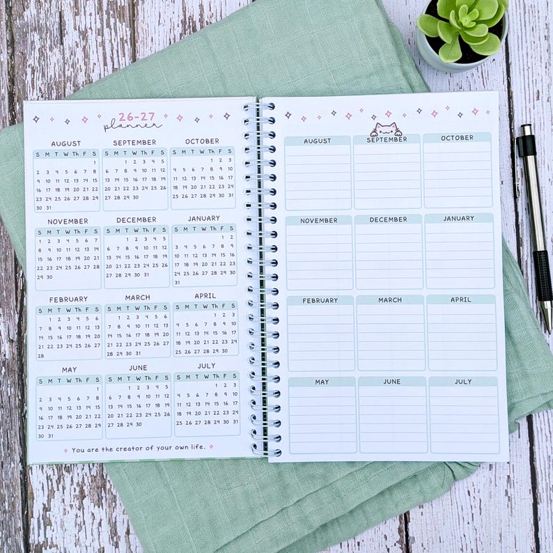 May include: A spiral-bound 26-27 planner with white pages featuring monthly calendar grids from August to July on the left and corresponding blank note sections on the right. The layout includes small star icons, a cat illustration, and the text 'You are the creator of your own life.'