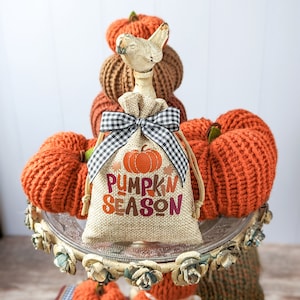 May include: A burlap bag with a black and white checkered ribbon and the text "Pumpkin Season" in a fall color palette. The bag is sitting on a tiered stand with crocheted pumpkins in orange and brown.