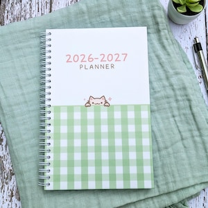 May include: A spiral-bound 2026-2027 planner with a white top section and a light green and white gingham patterned bottom section. The cover features the text 2026-2027 PLANNER in pink and brown, with a small illustration of a cat peeking over the gingham.