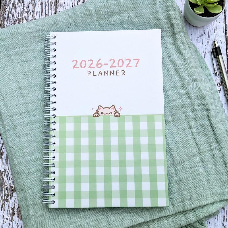 May include: A spiral-bound 2026-2027 planner with a white top section and a light green and white gingham patterned bottom section. The cover features the text 2026-2027 PLANNER in pink and brown, with a small illustration of a cat peeking over the gingham.