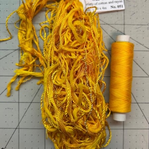 Pode incluir: Uma foto em close-up de linha e fio amarelos. Uma pilha emaranhada de fio amarelo está ao lado de um carretel de linha amarela. Uma etiqueta diz "One Offs" e "variety of cotton and rayon".