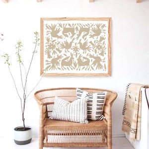 Op de afbeelding: Een ingelijste print van een beige volkskunstontwerp met gestileerde vogels, bloemen en dieren. De print hangt aan een witte muur boven een rieten stoel met zwart-witte gepatroneerde kussens.