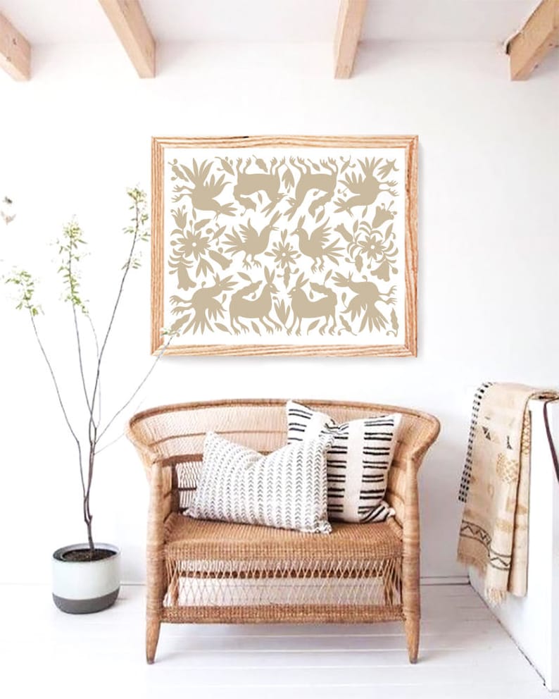 Op de afbeelding: Een ingelijste print van een beige volkskunstontwerp met gestileerde vogels, bloemen en dieren. De print hangt aan een witte muur boven een rieten stoel met zwart-witte gepatroneerde kussens.