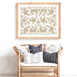 Op de afbeelding: Een ingelijst print met een beige ontwerp van gestileerde vogels, konijnen en bloemen op een witte achtergrond. De print hangt aan een witte muur boven een rieten bank met zwart-witte gepatroneerde kussens.