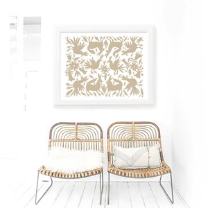 Op de afbeelding: Twee rieten stoelen met witte kussens staan voor een witte muur met een ingelijst afdruk van een beige volkskunstontwerp met vogels en konijnen.