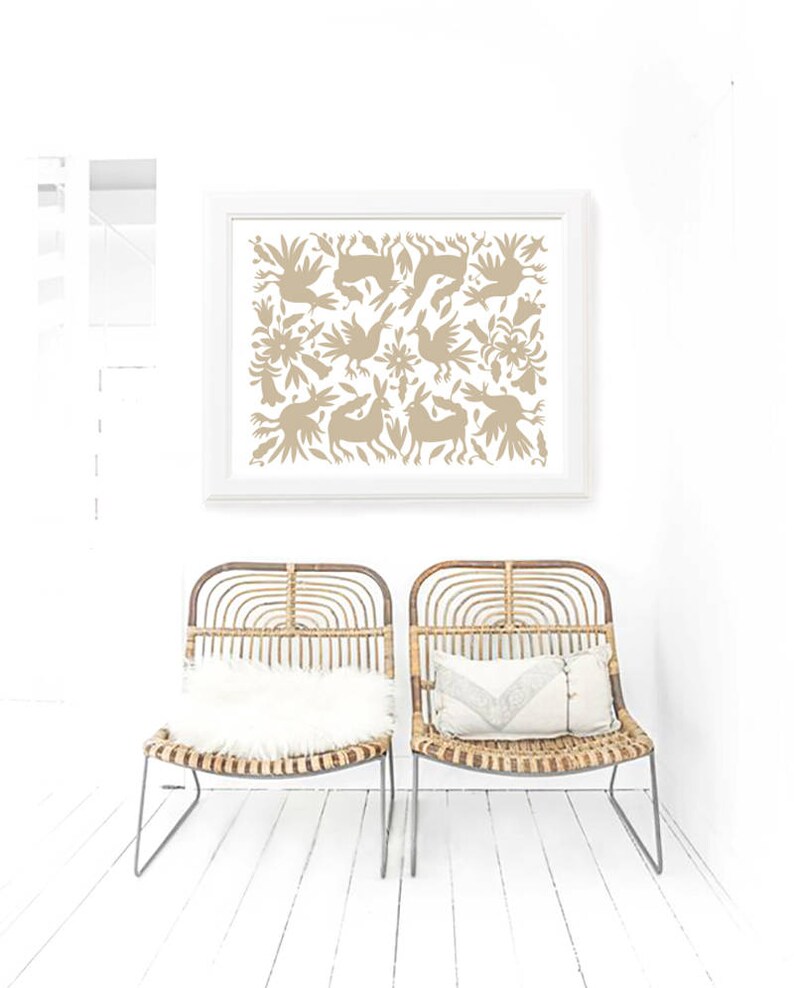Op de afbeelding: Twee rieten stoelen met witte kussens staan voor een witte muur met een ingelijst afdruk van een beige volkskunstontwerp met vogels en konijnen.