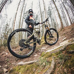 May include: An olive green mountain bike with black tires and suspension is positioned on a forest trail. The bike has a black seat and handlebars. The scene is set in a forest with tall trees and a person wearing a helmet and riding gear.