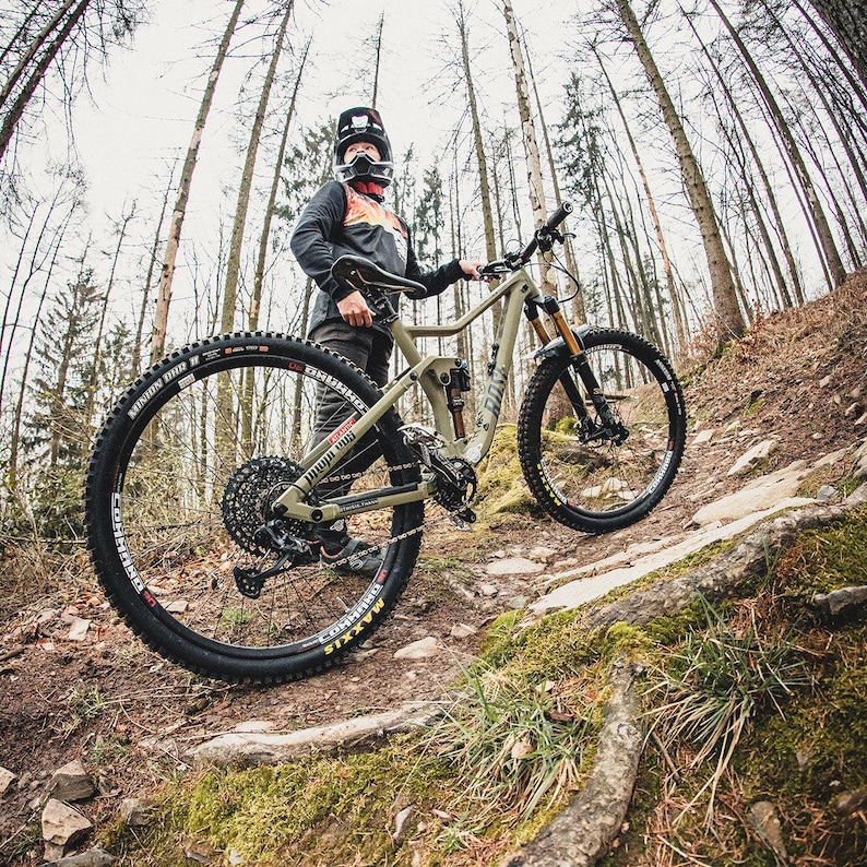 May include: An olive green mountain bike with black tires and suspension is positioned on a forest trail. The bike has a black seat and handlebars. The scene is set in a forest with tall trees and a person wearing a helmet and riding gear.