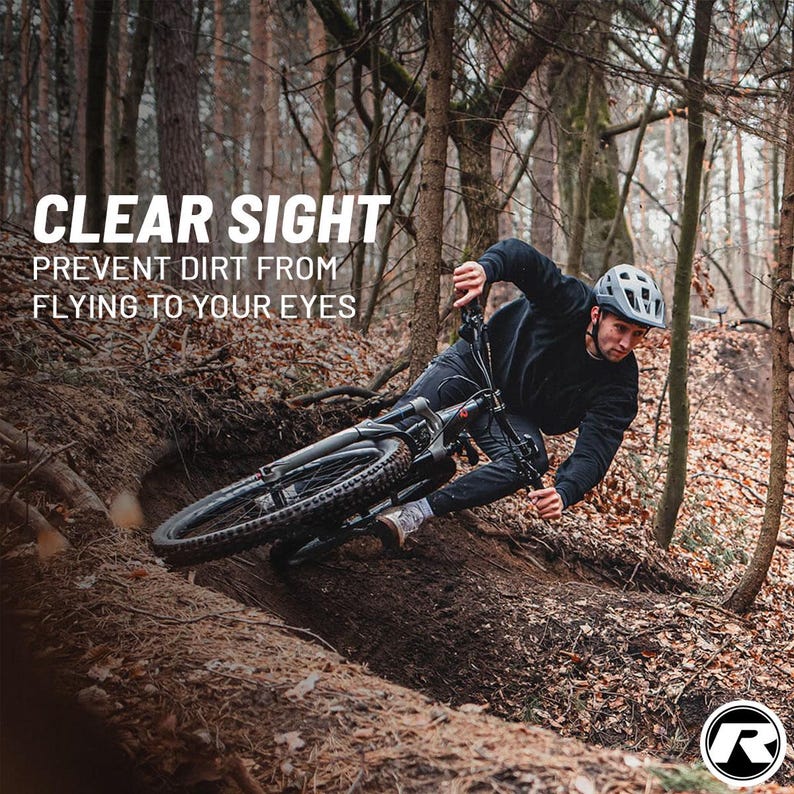 May include: A mountain biker navigates a forest trail on a black bicycle. The image includes the text "CLEAR SIGHT" and "PREVENT DIRT FROM FLYING TO YOUR EYES". The biker wears a helmet and dark clothing.