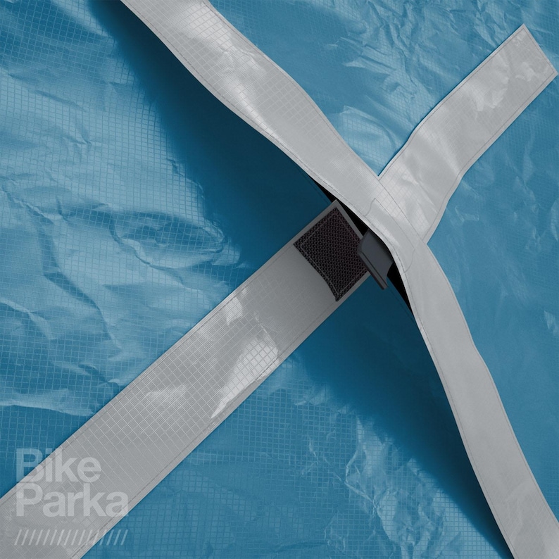 May include: Close-up of a blue Bike Parka cover with white straps crossing in an X shape. The straps have a grid pattern. The Bike Parka logo is visible in the lower left corner.