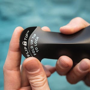 May include: A black tool with the text "FLIP TIP" and "FOLD UP TIP TO LOCK" printed in white. The tool is being held by two hands. The tool has a curved shape with a locking mechanism.