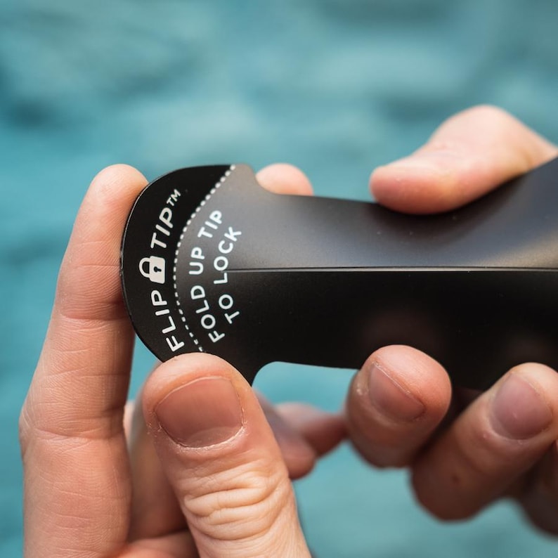 May include: A black tool with the text "FLIP TIP" and "FOLD UP TIP TO LOCK" printed in white. The tool is being held by two hands. The tool has a curved shape with a locking mechanism.