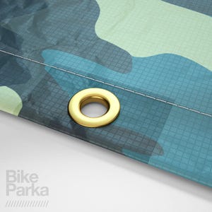 May include: Close-up of a section of a camouflage-patterned fabric in shades of blue and green. A gold-colored grommet is visible near the edge. The text "Bike Parka" is printed in white on the lower left corner.