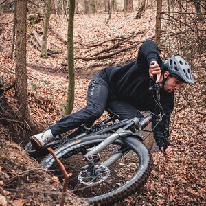 May include: A mountain biker in a black outfit and helmet navigates a forest trail. The black mountain bike has thick tires and disc brakes. The scene is set in a forest with fallen leaves on the ground.