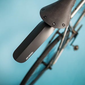 May include: Black bicycle fender with the text "ASS SAVERS" attached to a black bicycle seat. The bicycle wheel and frame are visible against a light blue background. The fender is designed to protect the rider from mud and water.