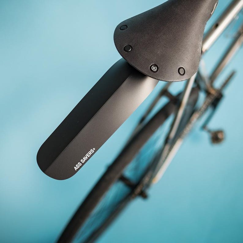 May include: Black bicycle fender with the text "ASS SAVERS" attached to a black bicycle seat. The bicycle wheel and frame are visible against a light blue background. The fender is designed to protect the rider from mud and water.