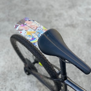 May include: A black bicycle seat with a colorful graphic mudguard. The mudguard features a collage of cartoon characters and designs. The bicycle tire is black with a yellow stripe. The seat post is black.