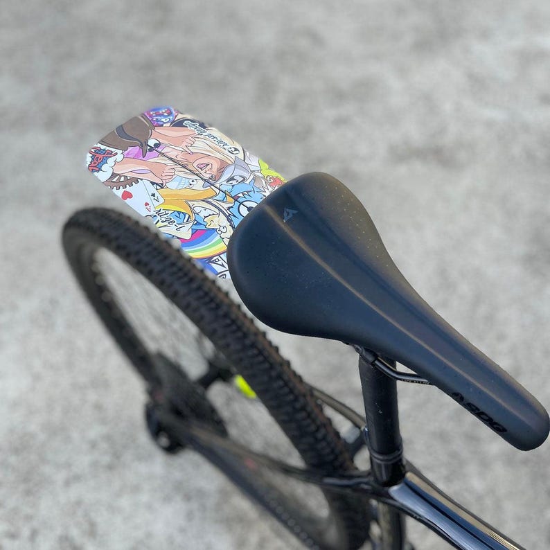 May include: A black bicycle seat with a colorful graphic mudguard. The mudguard features a collage of cartoon characters and designs. The bicycle tire is black with a yellow stripe. The seat post is black.
