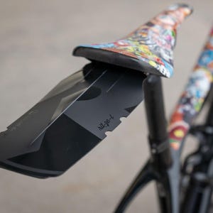 May include: A black bicycle mudguard with a geometric design and the text "rit-ze-l" is mounted on a bicycle. The bicycle seat has a colorful graphic design. The bicycle frame is black.