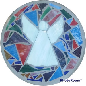 May include: A round, mosaic stepping stone featuring a white angel figure. The angel is surrounded by a colorful border of irregularly shaped glass pieces in shades of blue, red, green, and purple. The stone has a gray concrete border.