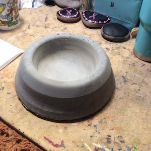 Peut inclure: Un bol en b&eacute;ton gris avec une base large et arrondie et un int&eacute;rieur peu profond. Le bol est sans ornements et repose sur une surface de travail. En arri&egrave;re-plan, des pierres peintes avec des rubans roses sugg&egrave;rent un th&egrave;me de soutien ou de sensibilisation.