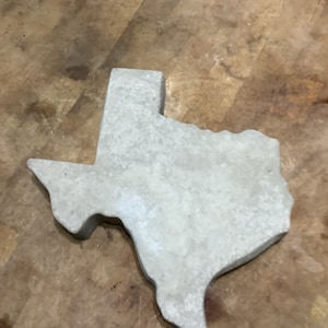 May include: A gray concrete Texas state outline. The outline is a three-dimensional shape and is sitting on a wooden surface.