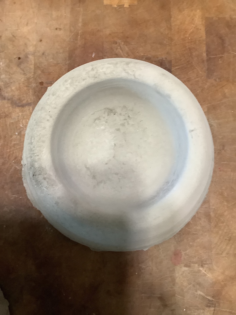 Peut inclure: Un mortier et pilon circulaires gris clair. Le mortier a un large bol peu profond avec une surface textur&eacute;e. Le pilon n'est pas visible. Le mortier est fait d'un mat&eacute;riau semblable &agrave; de la pierre et repose sur une surface en bois.