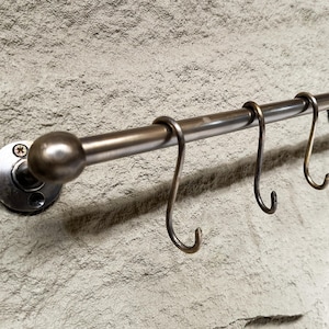 Könnte beinhalten: Eine horizontale Metallstange mit glatter, gebürsteter Oberfläche und abgerundeten Endkappen, die mit runden Halterungen an einer Wand befestigt ist. Drei S-förmige Metallhaken hängen an der Stange und dienen zur hängenden Aufbewahrung.