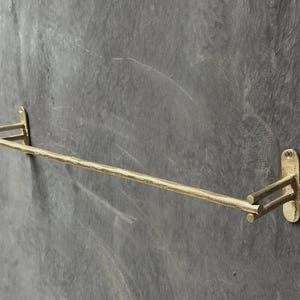 May include: A wall-mounted towel bar with a textured, metallic gold finish. The horizontal rod is supported by two double-arm brackets, each featuring a flat, oval-shaped mounting plate with a single screw hole at the top for secure installation.