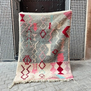Puede incluir: Una alfombra rectangular tejida a mano con un patrón geométrico. La alfombra presenta una base color crema con una sección central gris. Formas geométricas rosas, rojas y turquesas están dispersas por la superficie. La alfombra tiene un borde con flecos.