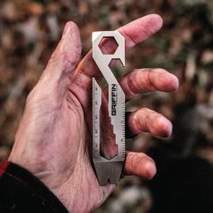 Könnte beinhalten: Ein silbernes Griffin Pocket Tool Multi-Tool. Das Werkzeug hat eine sechseckige Öffnung, einen Flaschenöffner, ein Lineal mit Zentimetermessungen und den Markennamen "Griffin Pocket Tool".