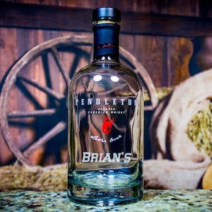 May include: A clear glass bottle of Pendleton Blended Canadian Whisky with a black label and a red logo of a horse's head. The bottle is sitting on a brown surface with a wooden wheel and a burlap sack in the background. The bottle is labeled "Brian's".
