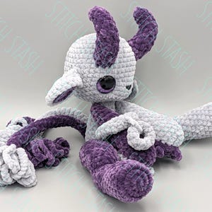 May include: A handmade crochet plush toy in shades of gray and purple. The plush features a round head with large black eyes, purple horns, and long, curling tentacles. The soft, textured yarn creates a whimsical and cuddly creature.