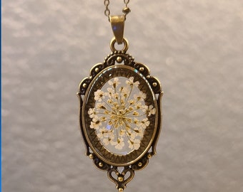 May include: A delicate white flower pressed inside a clear oval resin pendant, set in an ornate bronze frame. The pendant hangs from a bronze chain.