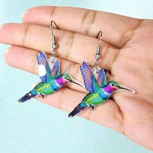 May include: A pair of hummingbird earrings. Each earring features a colorful hummingbird design with blue, green, purple, and pink hues. The earrings have silver-colored hooks.