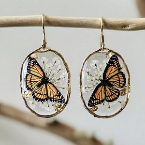May include: A pair of oval-shaped earrings featuring a butterfly design. The earrings have a gold-colored frame and a clear resin center with a butterfly and small white flowers. The butterfly has orange, black, and white markings.