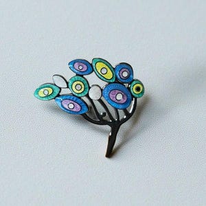 May include: A silver-toned brooch in the shape of a stylized tree. The brooch features colorful, oval-shaped enamel leaves in shades of blue, green, yellow, and purple. The leaves have white and purple centers. The brooch is a unique piece of jewelry.
