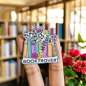 May include: A colorful enamel pin featuring a stack of books with flowers sprouting from the tops. The pin has the word "BOOKTROVERT" in white text on a black background. The books are in shades of orange, teal, and purple.