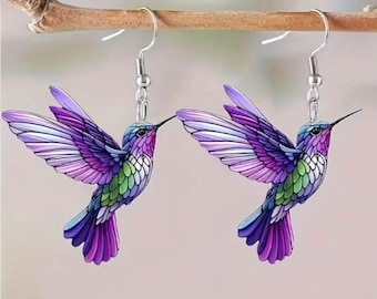 May include: A pair of hummingbird earrings. Each earring features a detailed, colorful hummingbird with outstretched wings. The birds are primarily purple, blue, and green, with silver-tone hooks. The earrings are suspended from a wooden branch.