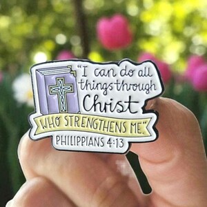 May include: A religious pin with the text "I can do all things through Christ who strengthens me" and "Philippians 4:13". The pin features a purple book with a gold cross and a white banner.
