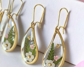 May include: Gold teardrop earrings with a clear resin filled with pressed flowers and greenery. The flowers are white and pink.