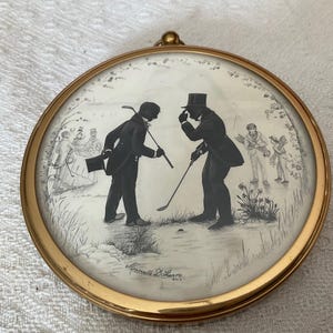 May include: A circular, gold-coloured framed artwork showing a golf scene in silhouette. Two men are playing golf, with other figures in the background. The artwork is in monochrome, with fine details and a vintage style.